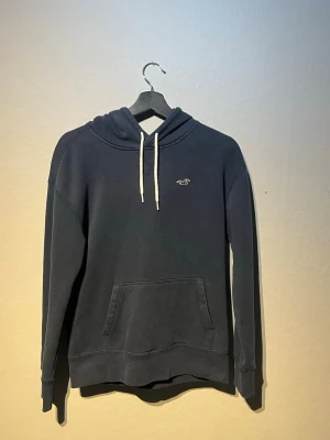 Hollister hoddie - Mörkblå hoodie från Hollister med vit dragsko i huvan och klassisk känguruficka framtill. Liten broderad logga på bröstet. Tillverkad i mjukt bomullsmaterial och har ribbade muddar vid ärmslut och nederkant.