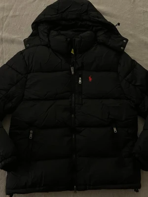 Svart Ralph Lauren Jacka - Svart dunjacka från Polo Ralph Lauren med huva och dragkedja framtill. Jackan har två sidofickor med dragkedja och den klassiska röda loggan på bröstet. Finns 1 innerficka samt avtagbar luva Perfekt för kalla dagar och har en quiltad design som ger en schysst look. Priset är ej hugget i sten. Köpt från Vinted men ångrar det och kan inte lämna tbx /mvh