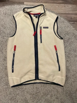 Vit fleeceväst från Patagonia - Säljer en vit fleeceväst från Patagonia med marinblå detaljer och röda dragkedjor. Västen har tre fickor med dragkedja och ett broderat Patagonia-märke på bröstet. Perfekt för lager-på-lager och outdoor-häng.