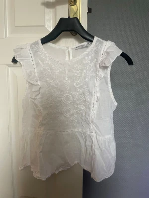 Vit broderad blus från Stradivarius - Söt vit blus från Stradivarius med broderade blommor framtill. Blusen är ärmlös med volangdetaljer vid axlarna och har en rund halsringning. Tillverkad i lätt bomull, perfekt för varma dagar. 