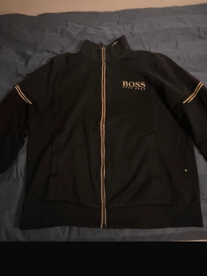 Hugo Boss helzip tröja  - Svart ziptröja från Hugo Boss med gulddetaljer längs dragkedjan och ärmarna. Tröjan har hög krage med logotyp på insidan och BOSS-brodyr på bröstet. Perfekt för en stilren och sportig look. Billig och är i storlek L