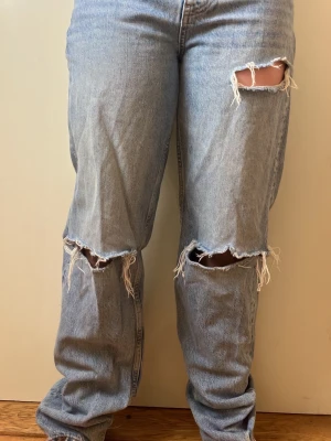 Ljusblå slitna straight jeans - Säljer ett par ljusblå jeans med raka ben och flera slitningar på både fram- och baksida. Jeansen har klassisk femficksdesign och hög midja. Perfekta för en avslappnad och trendig look. Har inte kommit kommit till användning 