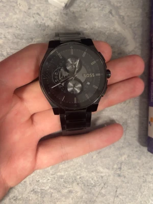 Svart kronografklocka från BOSS - Stilren svart kronografklocka från BOSS med metallarmband och flera urtavlor. Klockan har en modern och elegant design med diskreta detaljer och logotyp på urtavlan. Perfekt accessoar för dig som gillar en klassisk men ändå trendig look.