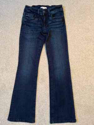 Bootcut jeans med låg midja från Gina Tricot Young, nyskick. - Snygga mörkblå bootcut jeans från Gina Tricot Young med klassisk femficksdesign och dragkedja. Jeansen har låg justerbar midja och är tillverkade i stretchigt denim för en bekväm passform. Perfekta för en trendig och avslappnad look. Knappt använda, nyskick.