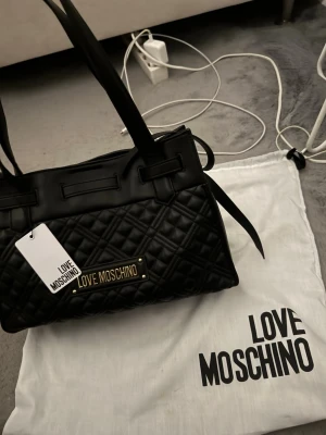 Svart quiltad handväska Love Moschino - Snygg svart handväska från Love Moschino med quiltad design och guldfärgad logga framtill. Väskan har dubbla handtag och detaljer i skinnimitation. Perfekt för dig som vill ha en trendig och rymlig väska med exklusiv känsla. Köpt i MILANO för 2500kr