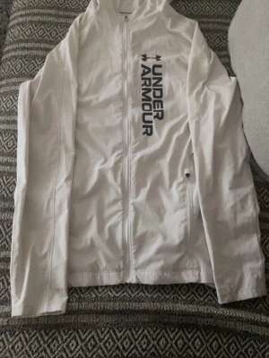 Vit vindjacka från Under Armour - Snygg vit vindjacka från Under Armour med huva och dragkedja framtill. Jackan har en stor logga i svart på bröstet och praktiska fickor med dragkedja. Perfekt för blåsiga dagar och sportiga outfits.