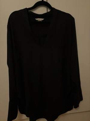 Svart v-ringad blus - Stilren svart blus med v-ringning och långa ärmar. Blusen har en lös passform och breda manschetter med knappdetalj. Tillverkad i ett mjukt, lätt material som ger ett snyggt fall. Perfekt att matcha med jeans eller kjol för en trendig look.