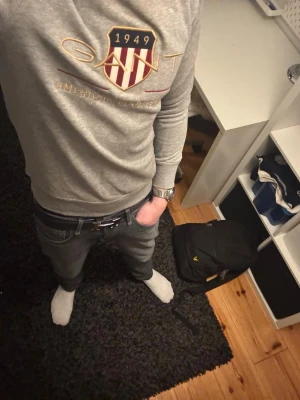 Grå sweatshirt från GANT - Säljer en grå sweatshirt från GANT med broderad logga och sköld på bröstet. Tröjan har texten 'American Sportswear' i guld och rött, rund halsringning och mjukt material. Perfekt för en avslappnad och stilren look.