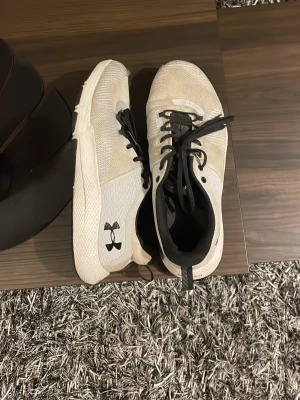 Vita sneakers från Under Armour - Snygga vita sneakers från Under Armour med svart logga på sidan och svarta snören. Ovandelen är i mesh-liknande syntetmaterial för bra andningsförmåga och komfort. Platt sula och låg profil gör dem perfekta för en sportig look. Lite skavda i hälarna