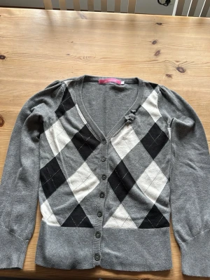 Grå argyle kofta från 365 Sunshine - Stilren grå kofta med argylemönster i svart och vitt och silvertråd från 365 Sunshine. V-ringad med knappar framtill och en söt liten rosett vid halsringningen. Långa ärmar och ribbade muddar. Perfekt för dig som gillar klassisk preppy stil. Storlek S. Skulle säga en liten s eller kan passa en son är xs 