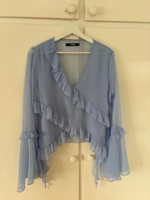 Ljusblå volangblus från BikBok - Supersöt ljusblå blus från BikBok med volangdetaljer och v-ringning. Blusen har transparent, lätt material och vida, långa ärmar med volanger. Storlek M🩷