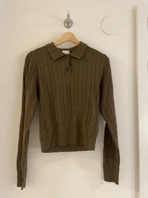 Olivgrön kabelstickad tröja med krage - Snygg olivgrön kabelstickad tröja med klassisk krage och två knappar framtill. Tröjan har långa ärmar och ribbade muddar. 