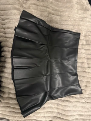 Svart skinnkjol - Snygg svart kortkjol i skinnimitation med plisserad design. Kjolen har en cool och edgy look som passar perfekt till många olika stilar. Storlek S och är använd en gång så kvaliteten är som ny!