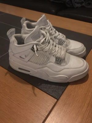Vita Nike Jordan 4 sneakers - Snygga helvita Nike Jordan 4 sneakers med klassisk siluett och meshdetaljer på sidorna. Skorna har snörning, rund tå och platt sula. Tillverkade i syntetmaterial och skinn för en clean och stilren look.