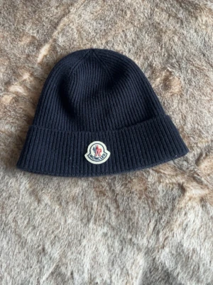 Blå  mössa från Moncler - Snygg blå ribbstickad mössa från Moncler med klassisk uppvikt kant och ikoniska Moncler-loggan framtill. Perfekt accessoar för kalla dagar och ger en clean look till din outfit.