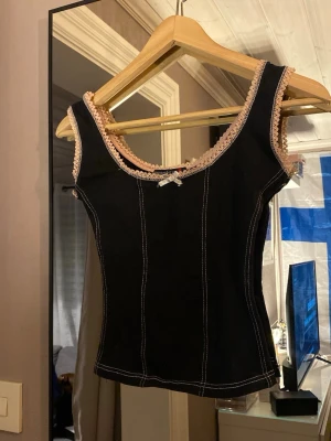 Svart topp med spets och rosett - Snygg svart topp från Brooker med ljusrosa spetsdetaljer runt halsringning och ärmhål. Dekorerad med en liten rosett framtill och kontrastsömmar som ger en cool look. Figurnära passform och ärmlös design, perfekt för att lyfta vilken outfit som helst.