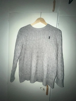 Ljusgrå kabelstickad tröja Polo Ralph Lauren - Ljusgrå kabelstickad tröja från Polo Ralph Lauren med rund halsringning och den klassiska broderade loggan på bröstet. Tröjan har långa ärmar och ribbade muddar vid ärmslut och nederkant. Perfekt för lager på lager och en clean look passar även S.