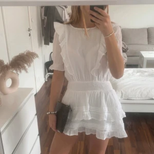 Vit student/skolavslutningsklänning - Supersöt vit volangklänning från Gina Tricot med volangdetaljer och smock i midjan. Klänningen har korta ärmar, rund halsringning och flera lager volanger på kjolen för en luftig känsla. Perfekt för sommarens alla tillfällen!