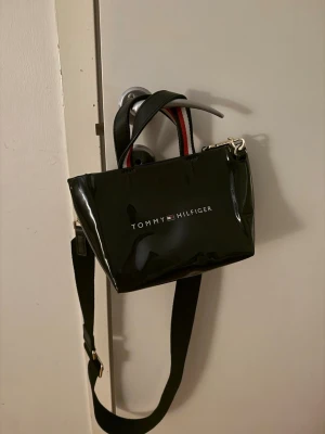 Svart handväska Tommy Hilfiger - Snygg svart handväska från Tommy Hilfiger med glansig finish. Väskan har både handtag och justerbar axelrem samt logga framtill. Handtagen har en cool röd och vit detalj som ger väskan en sportig vibe. Perfekt för dig som vill ha en stilren och trendig accessoar.