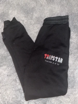 Svarta Trapstar mjukisbyxor - Svarta mjukisbyxor från Trapstar med trycket 'Trapstar It's a Secret' i vitt och rött på benet. Byxorna har resår i midjan och muddar vid bensluten. Perfekta för chill eller streetwear-stil.