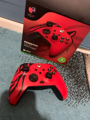 Xbox Kontroll - Snygg röd PDP Rematch Spirit Red handkontroll till Xbox med svart mönster och svarta knappar. Ergonomisk form med greppvänliga handtag och klassisk Xbox-layout. Tillverkad i plast med matta och blanka detaljer. Perfekt för gaming och snygg design som sticker ut.