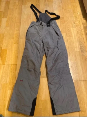 Grå täckbyxor med hängslen - Säljer ett par grå täckbyxor från Mckinley med justerbara svarta hängslen och knäppning fram. Byxorna har förstärkta partier vid bensluten och praktiska fickor. Perfekta för kalla dagar och vinteraktiviteter.