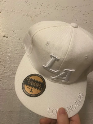 Vit LA snapback från Leader - Snygg vit snapback-keps från Leader med broderad LA-logga framtill och texten 'Los Angeles' på skärmen. Klassisk rak skärm och justerbar baksida. Tillverkad i ett tåligt tygmaterial som ger en clean och modern look.