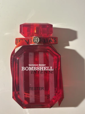 Bombshell Intense parfym Victoria's Secret - Bombshell Intense från Victoria's Secret i en snygg röd, multifacetterad glasflaska med dekorativ rosett och gulddetalj runt halsen. Flaskan har tydlig logga och text i vitt och svart. Perfekt för dig som gillar lyxiga och feminina dofter.
