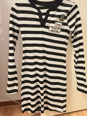 Randig svartvit klänning  - Säljer en svartvit randig klänning med lång ärm och ribbad struktur. Klänningen har V-ringning och coola patches på bröstet. Passformen är normal och längden är kort, perfekt för en avslappnad och snygg stil.