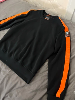 Svart sweatshirt från Parajumpers - OBS FLÄCKEN GÅR BORT MED VATTEN Cool svart sweatshirt från Parajumpers med orangea ränder längs ärmarna och ett broderat PJS-märke på axeln. Tröjan har rund halsringning och ribbade muddar. Perfekt för en sportig och avslappnad stil.