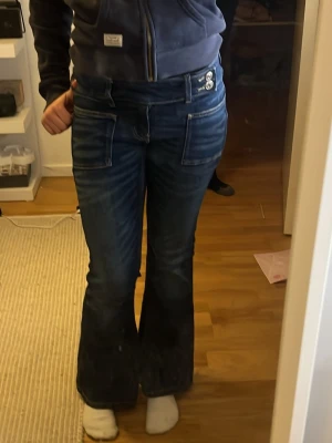 Blå bootcut jeans från Gina Tricot - Snygga blå bootcut jeans från Gina Tricot med dubbla knappar i midjan och stora fickor fram. Jeansen har en lågmidjad passform och är tillverkade i klassiskt denimtyg. Perfekta för dig som gillar en retroinspirerad look.