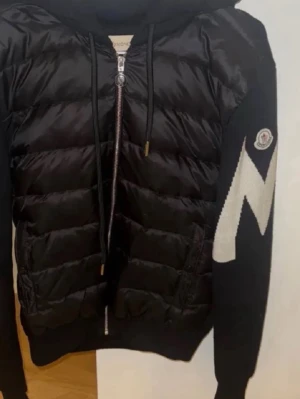 Svart cardigan jacka från Moncler - Snygg svart cardigan jacka från Moncler med quiltad framsida och stickade ärmar. Jackan har dragkedja, huva med snörning och ett stort vitt zigzag-mönster på ena ärmen samt Moncler-logga. Perfekt för dig som vill ha en sportig och trendig look.
