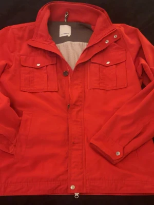 Röda J.lindeberg field jacket  - Snygg röd vindjacka med två bröstfickor med lock och knappar samt dragkedja framtill. Jackan har klassisk krage och långa ärmar. Perfekt för dig som vill sticka ut med färg och stil. Materialet känns lätt och smidigt.