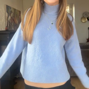 Ljusblå stickad polotröja - Mysig ljusblå stickad tröja med hög polokrage och bred ribbstickad kant nedtill samt vid ärmsluten. Tröjan har en avslappnad passform och är perfekt för kyliga dagar. Materialet ser mjukt och fluffigt ut, vilket ger en skön känsla. Den är dock nopprig, men går säkert att fixa en en noppmaskin!