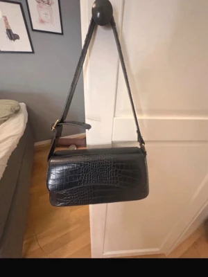 Svart axelremsväska i crocopräglat skinn - Snygg svart handväska med crocopräglat mönster och axelrem. Väskan har ett stilrent, avlångt format och innerfack med dragkedja. Perfekt för dig som vill ha en trendig accessoar med plats för det viktigaste.