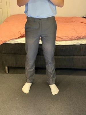 Grå kostymbyxor med rak passform - Snygga grå kostymbyxor med klassisk rak passform och pressveck. Byxorna har bälteshällor, knapp och dragkedja framtill. Perfekta för en stilren look och passar till skjorta eller t-shirt.