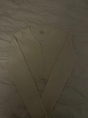 Vit ribbad kofta med knappar - Säljer en stilren vit ribbad kofta med v-ringning och långa ärmar. Koftan har en croppad passform och knäpps framtill med små vita knappar. Perfekt att matcha med jeans eller kjol för en enkel och fräsch look. Funkar som en storlek xss/xs också, då den är stretchig. 