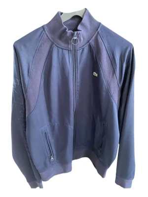 Mörkblå zip hoodie från Lacoste LIVE - Säljer en mörkblå hoodie från Lacoste LIVE i unisexmodell. Tröjan har hel dragkedja med rund zip puller, ståkrage och två fickor med dragkedja. Liten broderad krokodil på bröstet och diskret mönster längs ena ärmen. Perfekt för chill dagar.