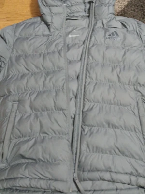 Grå dunjacka från Adidas med huva - Snygg grå dunjacka från Adidas med huva och dragkedja framtill. Jackan har quiltad design, två sidofickor och Adidas-logga på bröstet. Perfekt för kalla dagar och enkel att matcha med olika outfits.