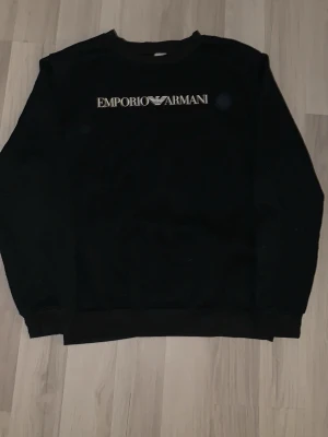 Svart sweatshirt från Emporio Armani - Stilren svart sweatshirt från Emporio Armani med logga och ikoniskt örntryck på bröstet. Tröjan har rund halsringning, långa ärmar och ribbade muddar vid ärmslut och nederkant. Perfekt för en clean och klassisk look.