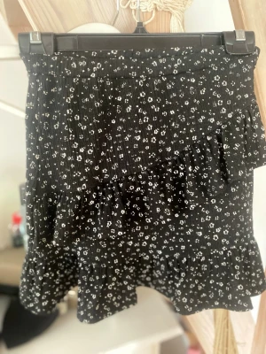 Svart blommig volangkjol - Kort svart kjol med små vita blommor och volangdetaljer. Kjolen har en feminin och lekfull look med flera lager volanger som ger extra rörelse. Perfekt för dig som gillar en söt och trendig stil.