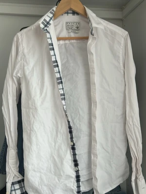 Vit skjorta med rutiga detaljer - Klassisk vit skjorta från Premium by Jack & Jones med rutigt mönster på insidan av kragen och knappslån. Skjortan har lång ärm, knappar framtill och är tillverkad i bomull. Perfekt för en clean och stilren look.