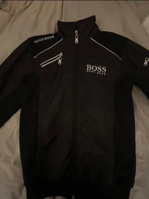 Svart vindjacka från Hugo Boss - Snygg svart vindjacka från Hugo Boss med vita detaljer och logga på bröstet och axeln. Jackan har hög krage, dragkedja framtill och en extra dragkedjeficka på bröstet. Perfekt för dig som vill ha en stilren och sportig look.