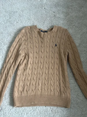 Beige kabelstickad tröja Polo Ralph Lauren - Kabelstickad beige tröja från Polo Ralph Lauren med rund halsringning och den klassiska mörkblå logotypen broderad på bröstet. Tröjan är i storlek M men jag skulle säga att den är lite större. (PRIS kan diskuteras vid snabb affär)