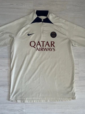 PSG beige träningströja med halv dragkedja - Snygg beige träningströja från PSG med halv dragkedja och mörkblå detaljer vid kragen. Tröjan har PSG-logga och Nike Swoosh på bröstet samt Qatar Airways-tryck framtill. Perfekt för fotbollsträning eller chill. Tillverkad i ett lätt och funktionellt material.