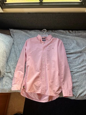 Rosa skjorta från Polo Ralph Lauren - Säljer en klassisk rosa skjorta från Polo Ralph Lauren i bomull. Skjortan har button-down krage, långa ärmar och den ikoniska broderade loggan på bröstet. Perfekt för en stilren och avslappnad look.