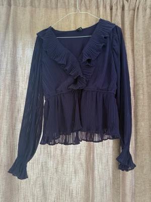 Mörklila plisserad blus Vero Moda - Snygg mörklila plisserad blus från Vero Moda med volangkantad omlott-ringning och långa transparenta ärmar. Blusen har ett band i midjan och lätt utsvängd nederdel. Perfekt för att lyfta vilken outfit som helst.