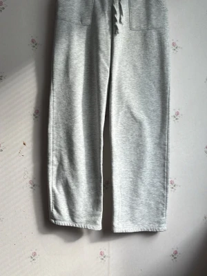 Ljusgrå mjukisbyxor med dragsko - Säljer ett par ljusgrå mjukisbyxor med bred resår i midjan och dragsko. Byxorna har raka ben och två stora fickor framtill. Tillverkade i mjukt sweatshirtmaterial som är perfekt för chill dagar. Passar dig som gillar en avslappnad och enkel stil.