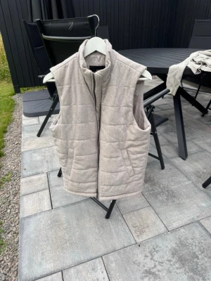 Beige dunväst från John Henric - Stilren beige dunväst från John Henric med hög krage och dragkedja framtill. Västen har quiltad design och två fickor på sidorna. Perfekt för lager-på-lager och passar till många olika outfits.