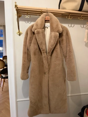 Beige fuskpäls-kappa Dry Lake - Säljer en beige lång fuskpäls-kappa från Dry Lake, modell Vanessa. Kappan har bred krage, rak passform och är helfodrad med ljus insida. Perfekt för att hålla värmen och samtidigt se riktigt stylish ut under kalla dagar.
